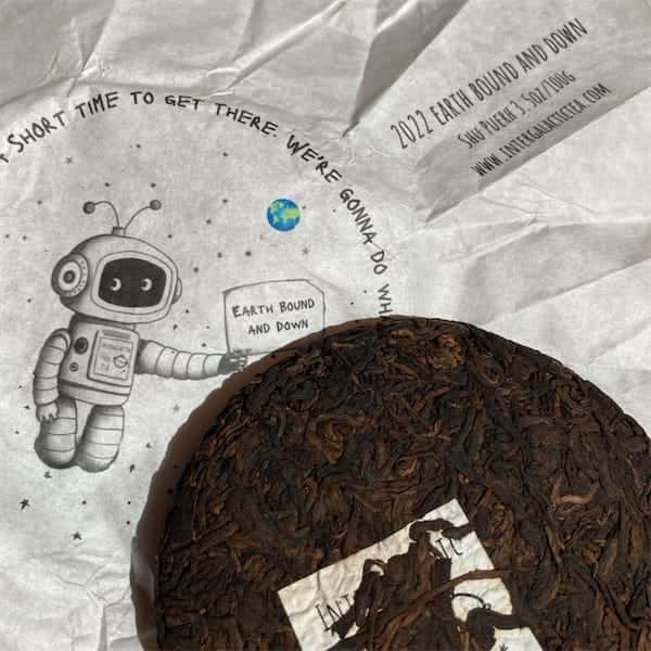 the compressed tea cake sitting on it's white paper wrapper, on the wrapper there is a cartoon like drawing of a robot spaceman