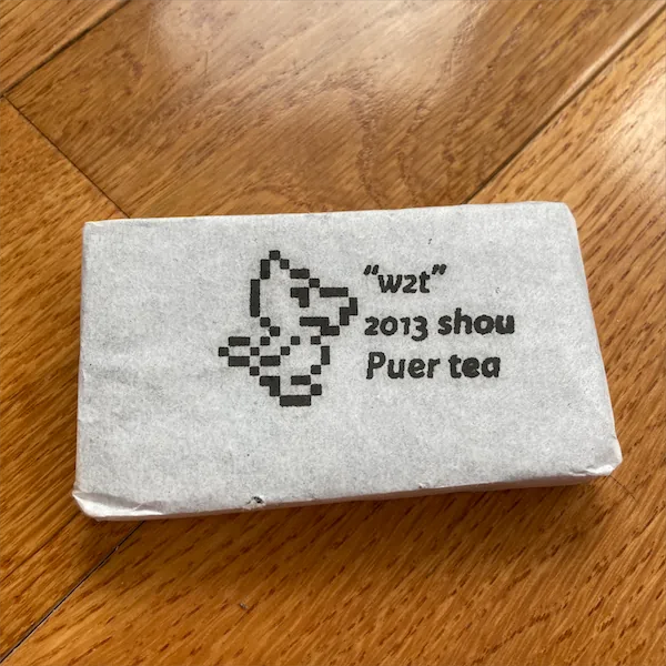 A white wrapped rectangle of tea sitting a wood floor. The wrapper has a pixelated image of a duck with the words w2t 2013 shou puer tea display next to the duck