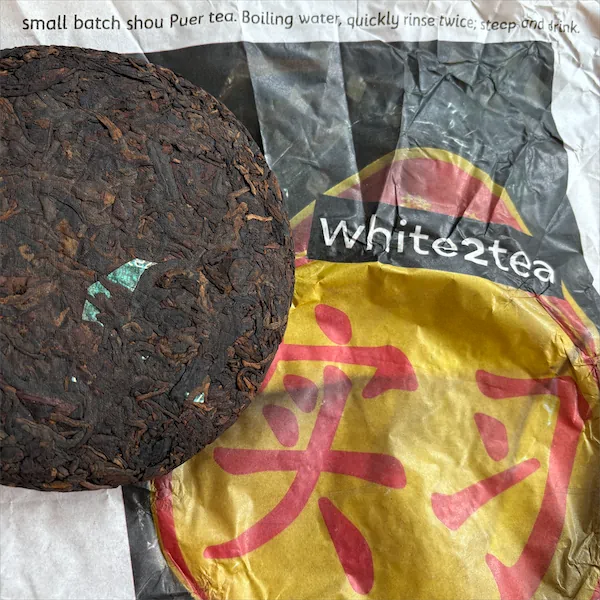 A dark brown mulch like looking tea cake sitting outside and on top of a yellow wrapper with red Chinese characters on it