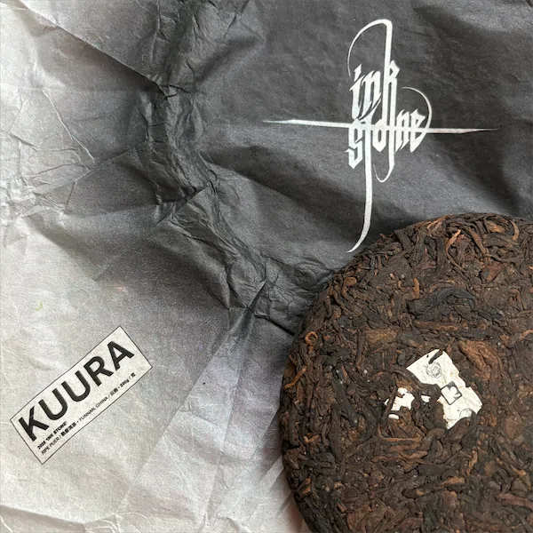 A tea cake wrapper that is a gradient of white to black with the Kuura logo and the Instone logos. On top of that sits the tea cake just off the edge of the image frame